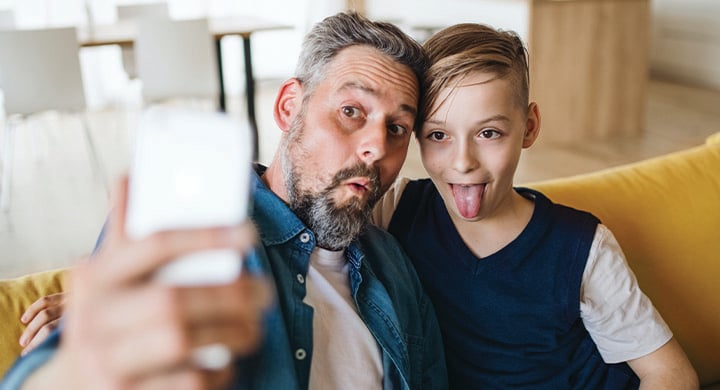 Mature father with small son sitting on sofa indoors, taking a funny selfie.