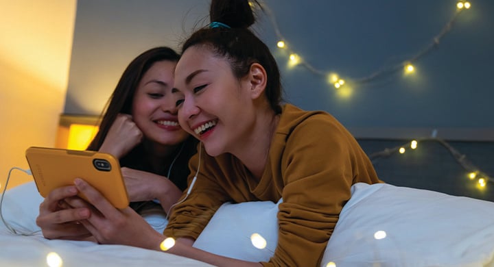  Asian woman friends lying on the bed in bedroom and watching movie or online shopping on smartphone together.