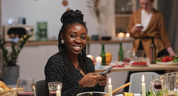 Portrait of young African woman smiling at camera while sitting at the table with her mobile phone and eating holiday dinner