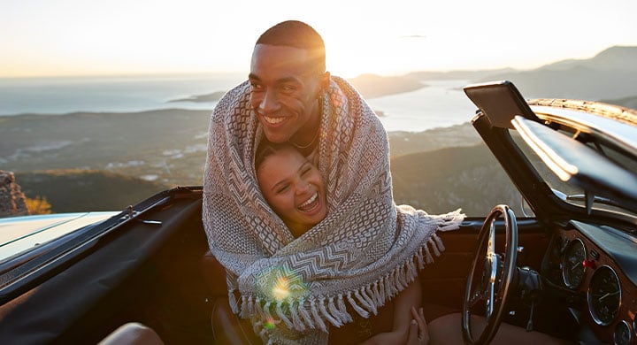 Happy man embracing girlfriend from behind while wrapped in blanket on road trip