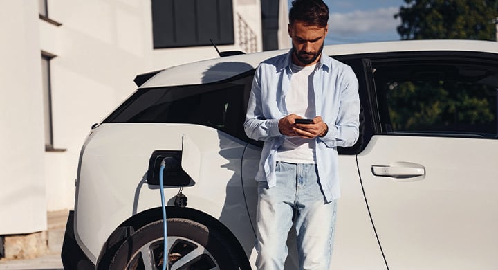 Sunny weather. Holding smartphone and charging the vehicle. Young stylish man is with electric car at daytime.
