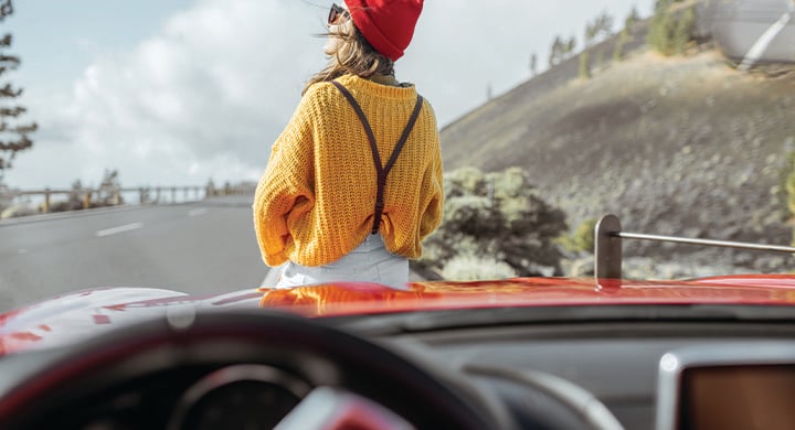 Lifestyle portrait of a carefree woman dressed casually in bright sweater and hat sitting on the car hood